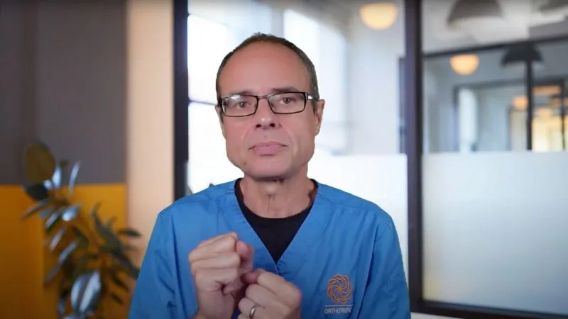 Dr. Mike Mew speaking to camera in blue clinical scrubs and glasses, hands raised in front of him, in a bright modern clinic.