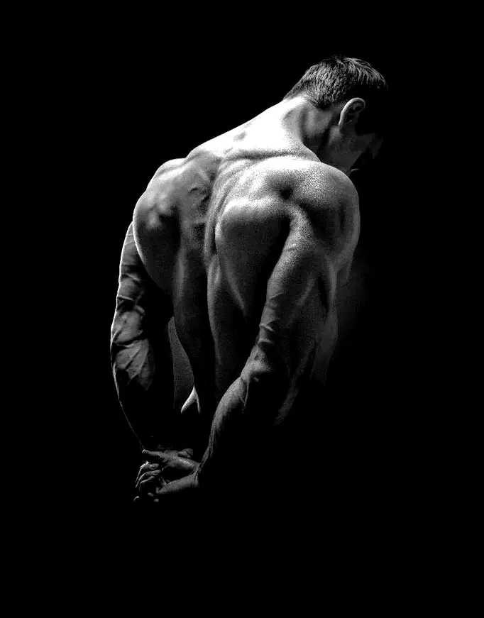 A muscular man's back and shoulders lit from one side against a black background, head lowered and hands clasped behind him.